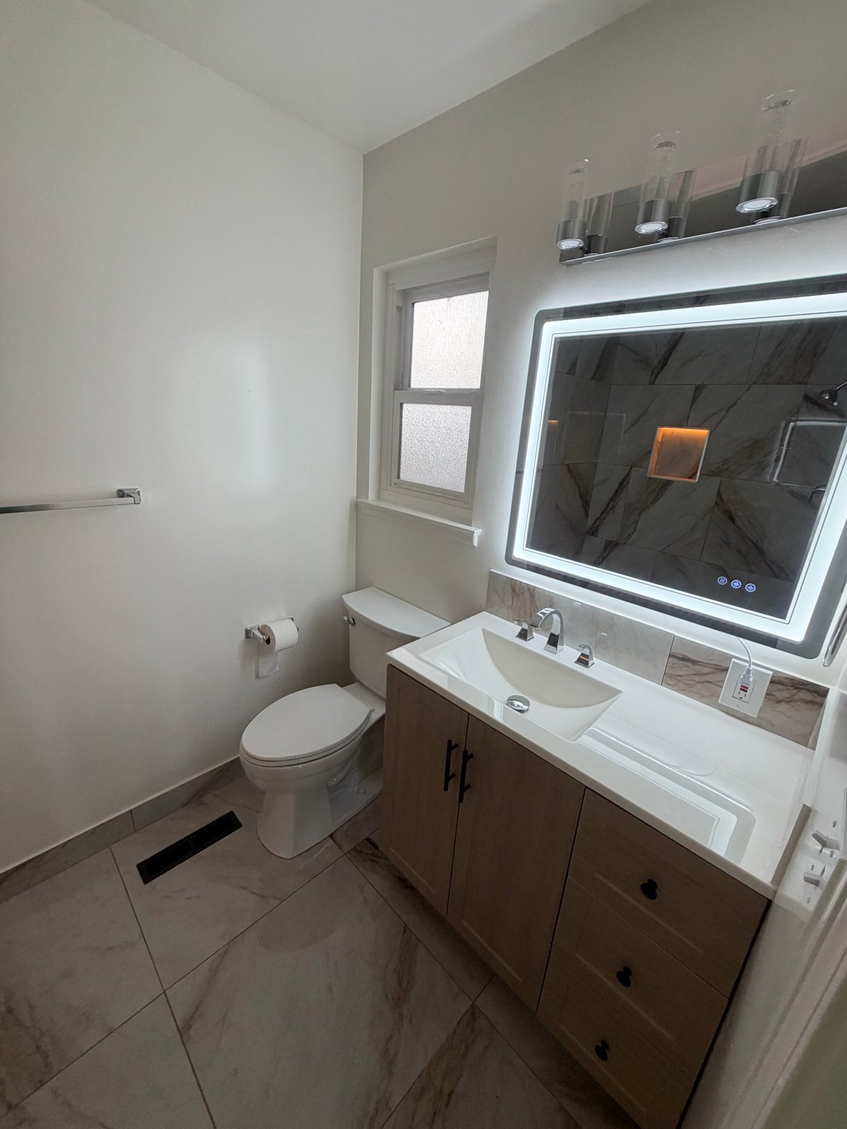 Modern bathroom with wood vanity, white sink, illuminated mirror, toilet, and window with frosted glass panes